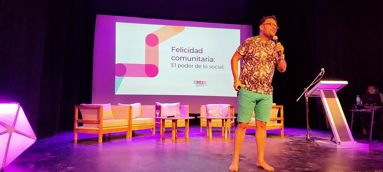 Un ponente en el escenario iluminado de rosa, con la pantalla «Felicidad comunitaria: el poder de lo social»